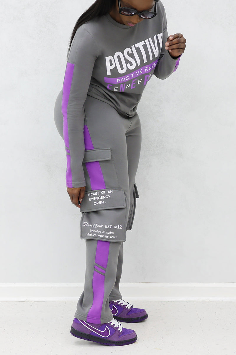 A woman wearing black sunglasses in a gray and purple tracksuit with pockets and purple shoes.