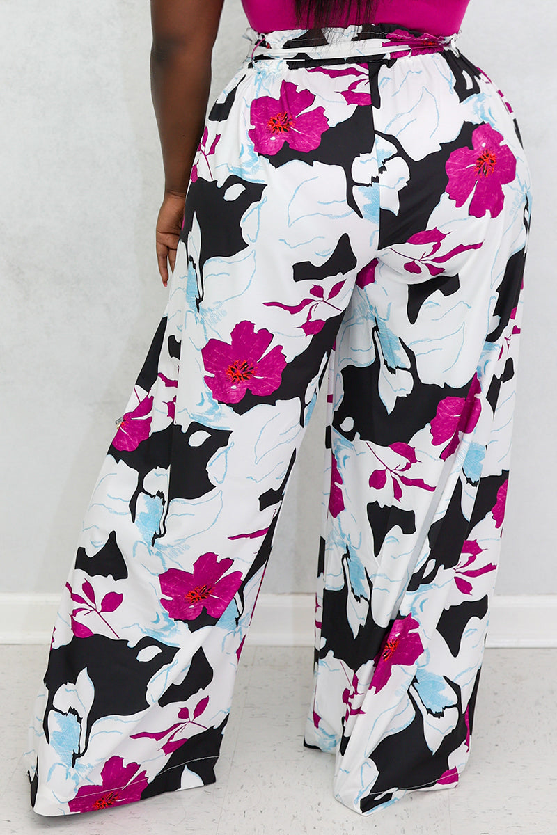 A person shown from the waist down stands indoors against a plain light-colored wall and floor, wearing Floral Palazzo Pants featuring a vibrant flowery pattern in pink, blue, black, and white.