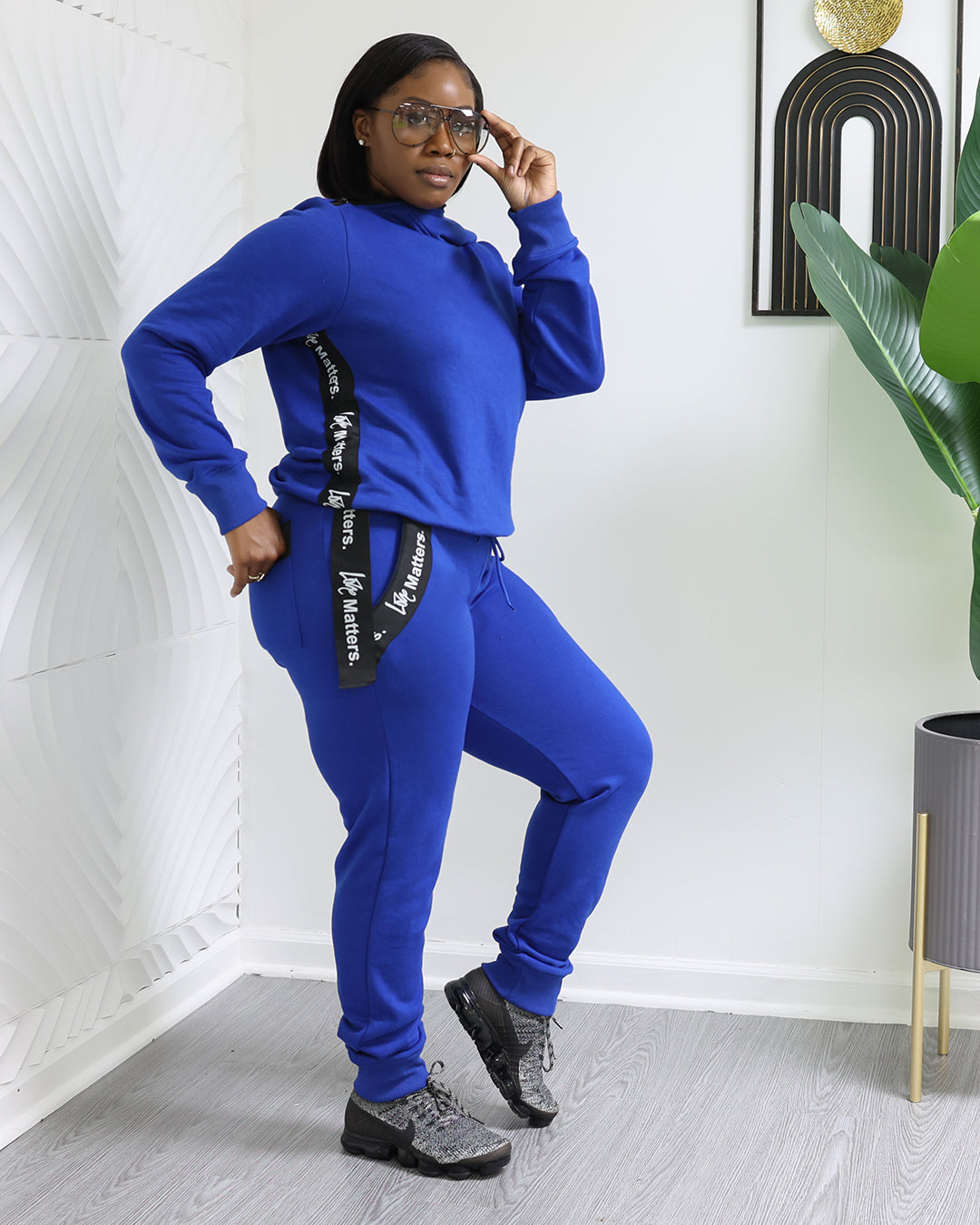 A confident individual wears Love Matters Joggers in a trendy blue tracksuit with black and white text on the sleeves, paired with black sneakers. They stand in a modern space adorned with geometric wall patterns, a green plant, and framed artwork.