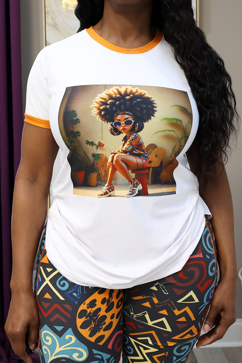 Wearing the Ife Binta T-Shirt in multi, featuring orange trim and an African American woman with a large afro and sunglasses, paired with colorful patterned leggings. The comfy plus-size tee offers 4-way stretch. The models face is not visible.