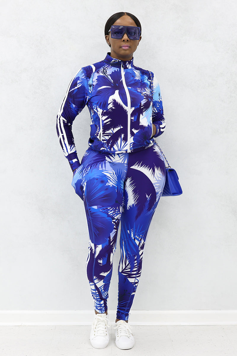 A woman wearing long-sleeved zipper jacket with blue and white tropical print, sunglasses, purse and white shoes.