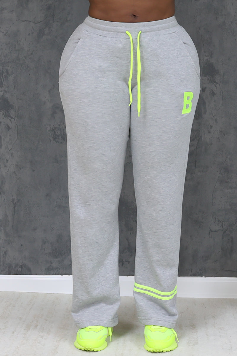 Wearing the Lime and Gray Leisure Pants in gray-green with neon yellow drawstrings and a B on the left leg, paired with matching neon yellow sneakers, a person stands before a textured gray wall.
