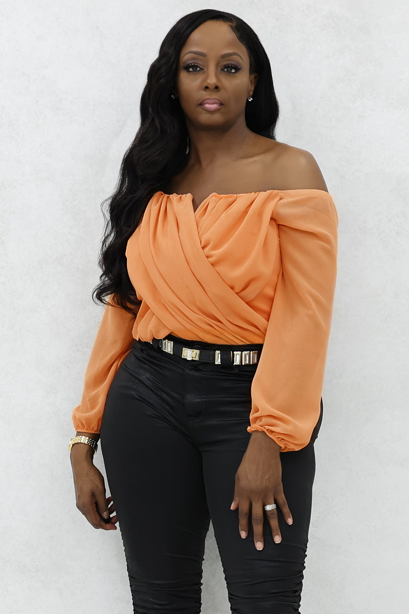 A woman wearing an off-shoulder peach top, black pants, and a gold watch.