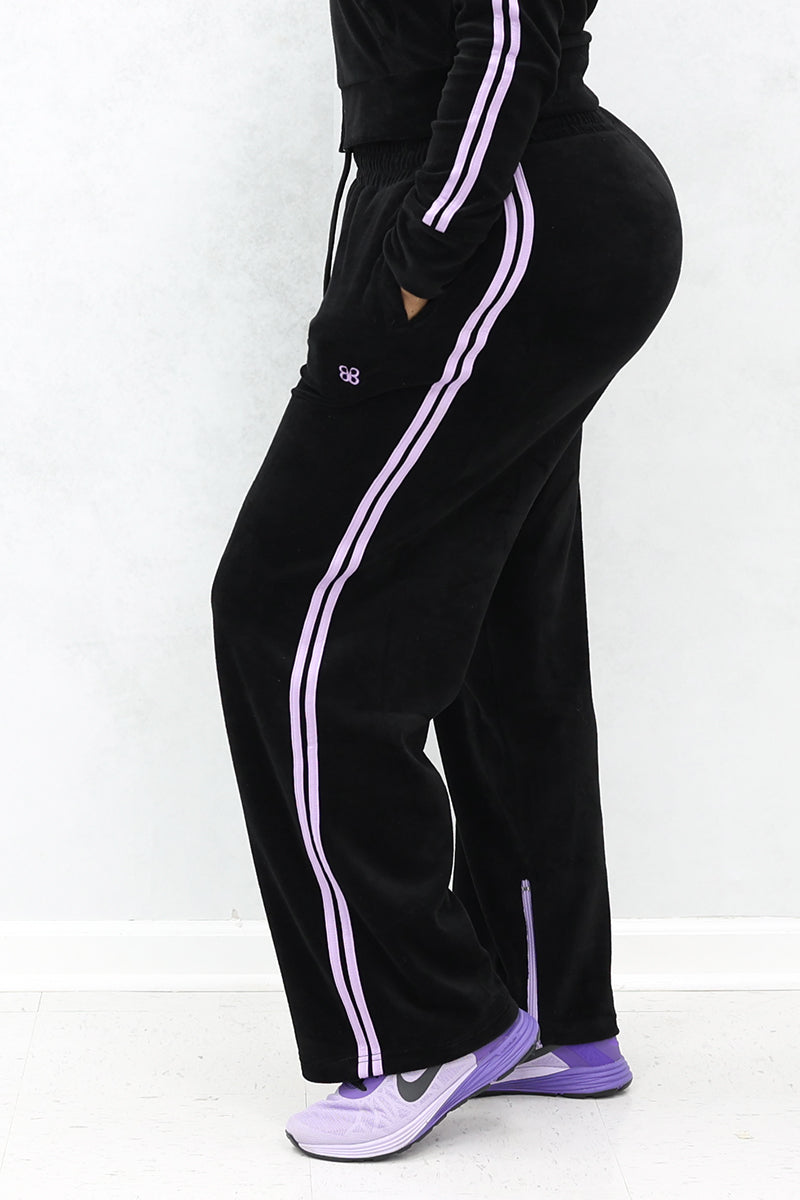 Black velour sweatpants with two purple side stripes. Model is wearing purple nike sneakers.