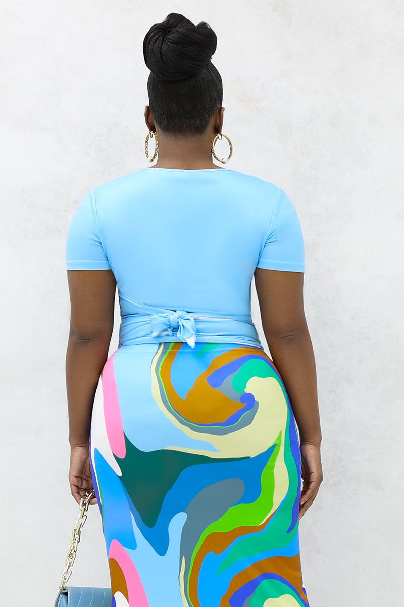 A woman wearing blue wrap tie top and colorful abstract print skirt holding a blue bag.