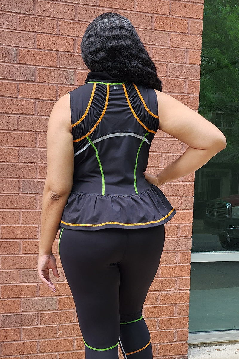 A person stands facing a brick wall, wearing the Ruffle Athleisure Jacket featuring a sleeveless black design with green and orange contrast stitching, a peplum hem, and matching black leggings. Their long hair falls in waves.