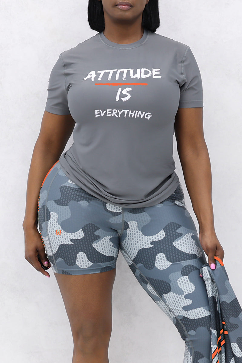 A person in a gray Attitude Is Everything T-shirt and blue-gray camo shorts, made from soft, lightweight fabric, holds a matching Polyester/Spandex camo garment against a plain light background.