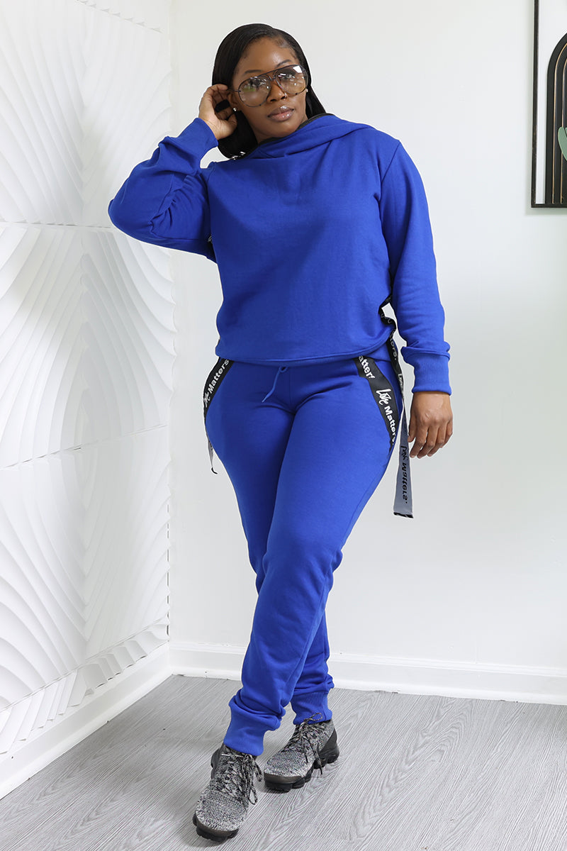A person in a bright blue tracksuit and gray sneakers stands indoors, adjusting their glasses. The room has a white textured wall and light gray floor. Wearing Love Matters Joggers with zippered details, they effortlessly blend fashion and function.