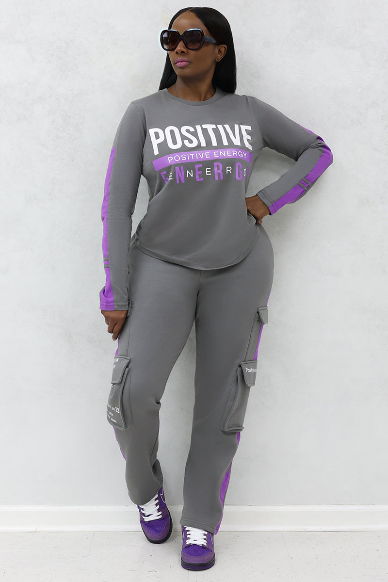 A woman wearing black sunglasses in a gray and purple tracksuit and purple shoes.