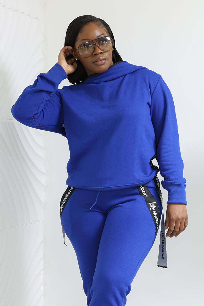 A person confidently poses against a textured white wall, wearing a cozy Love Matters Hoodie and matching breathable pants with texted side stripes, complemented by large sunglasses.