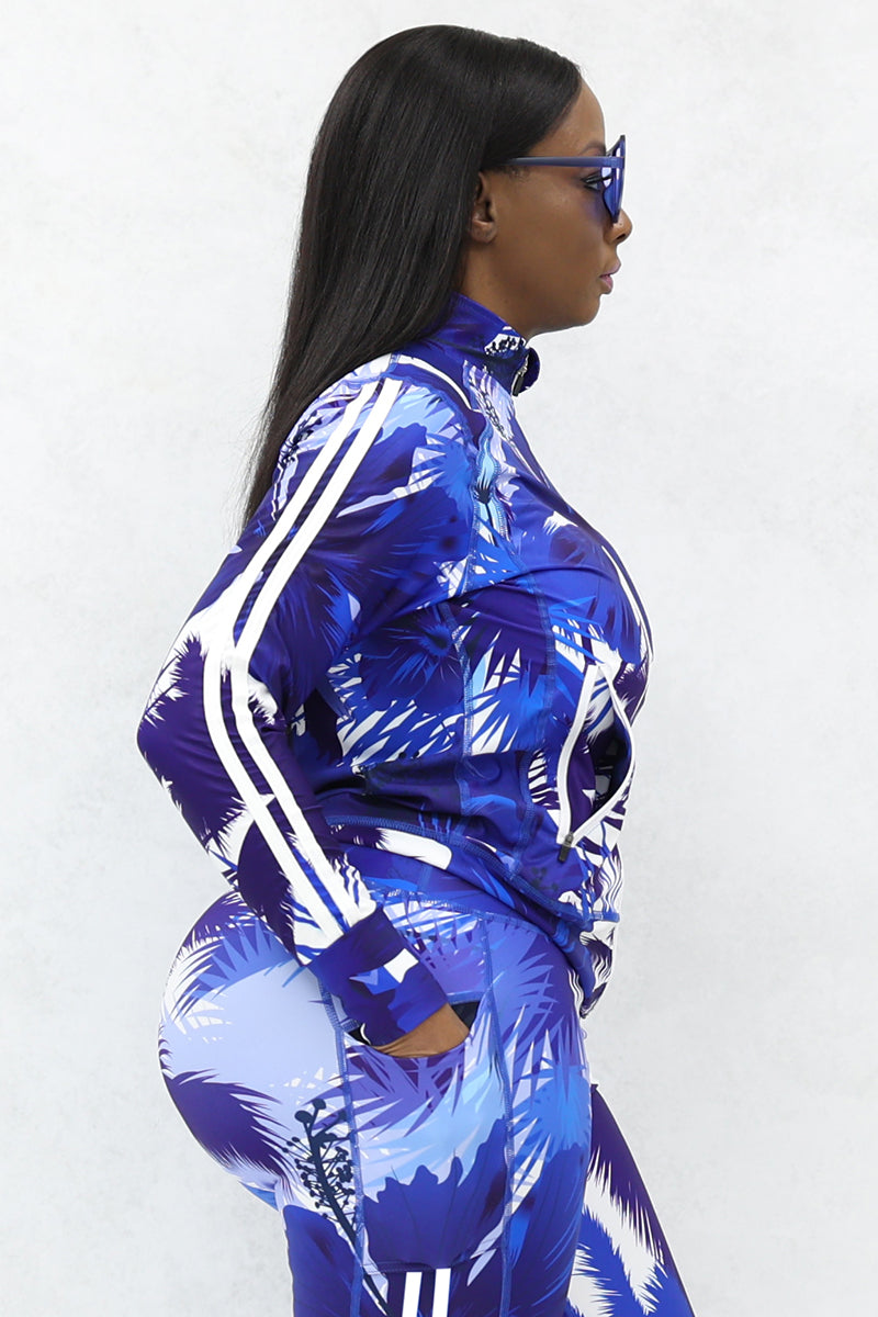 A woman wearing blue sunglasses, long-sleeved zipper jacket with blue and white tropical print and pockets.