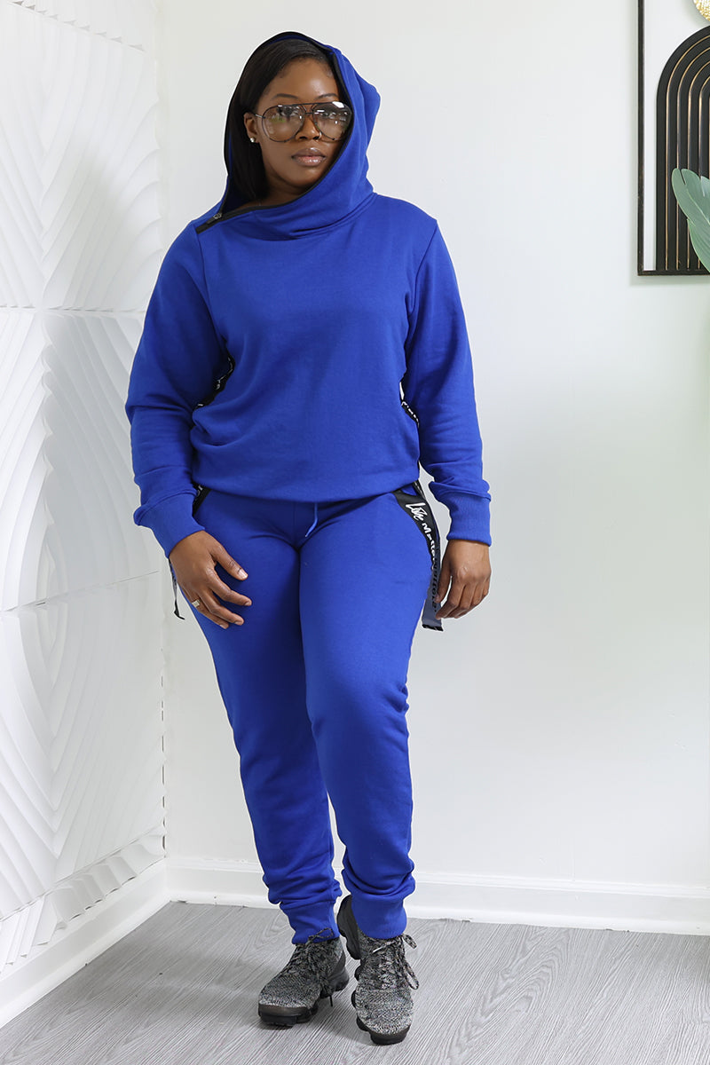 Someone wearing Love Matters Joggers poses indoors against a white patterned wall, dressed in a royal blue tracksuit with a hood. They stand on a light-colored floor, sporting glasses and black sneakers. A framed artwork and small plant are partially visible in the background.
