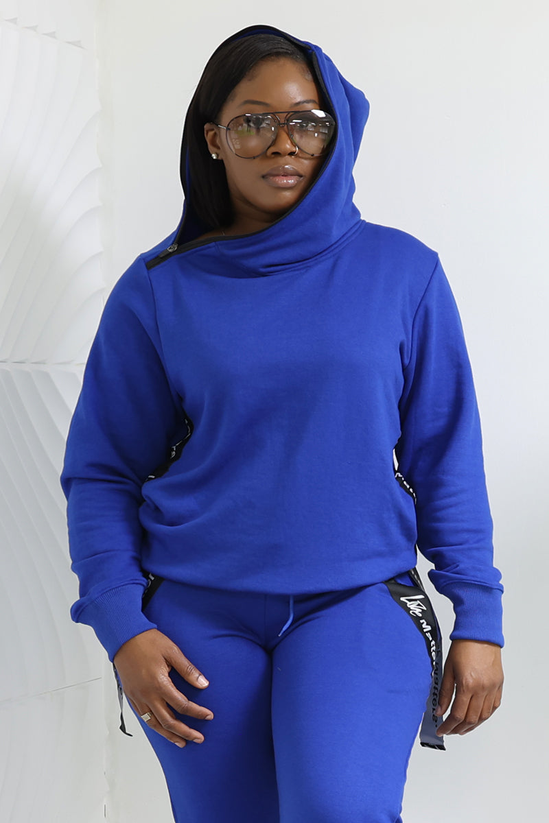 A person in a royal blue Love Matters Hoodie made from breathable fabric, hood up, wearing matching pants. With long dark hair and large round glasses, they exude cozy comfort against a neutral white background.