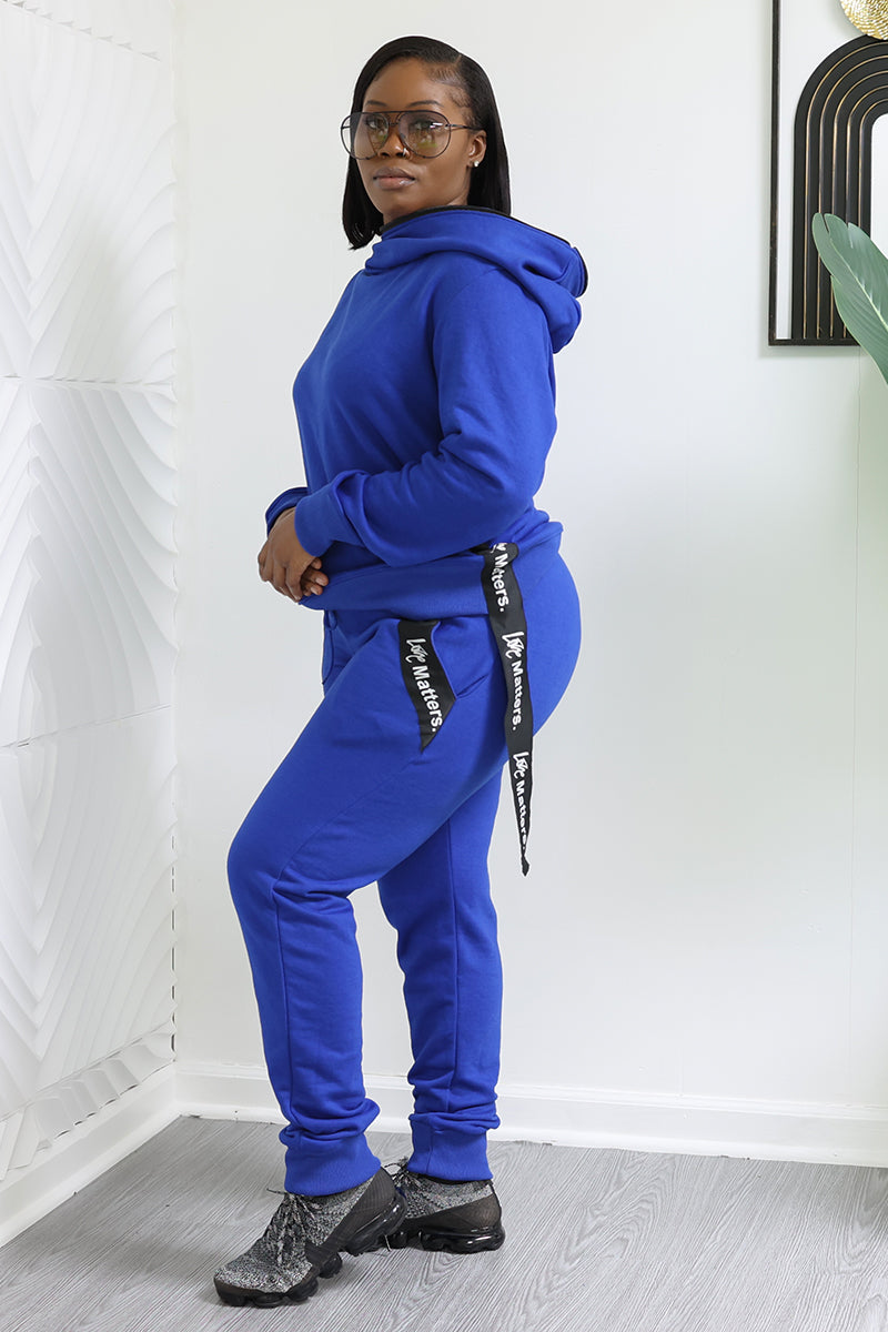 Indoors, a person poses in a blue hoodie and Love Matters Joggers with black ribbon accents and white text. They wear glasses and black shoes, set against white walls with decorative elements.