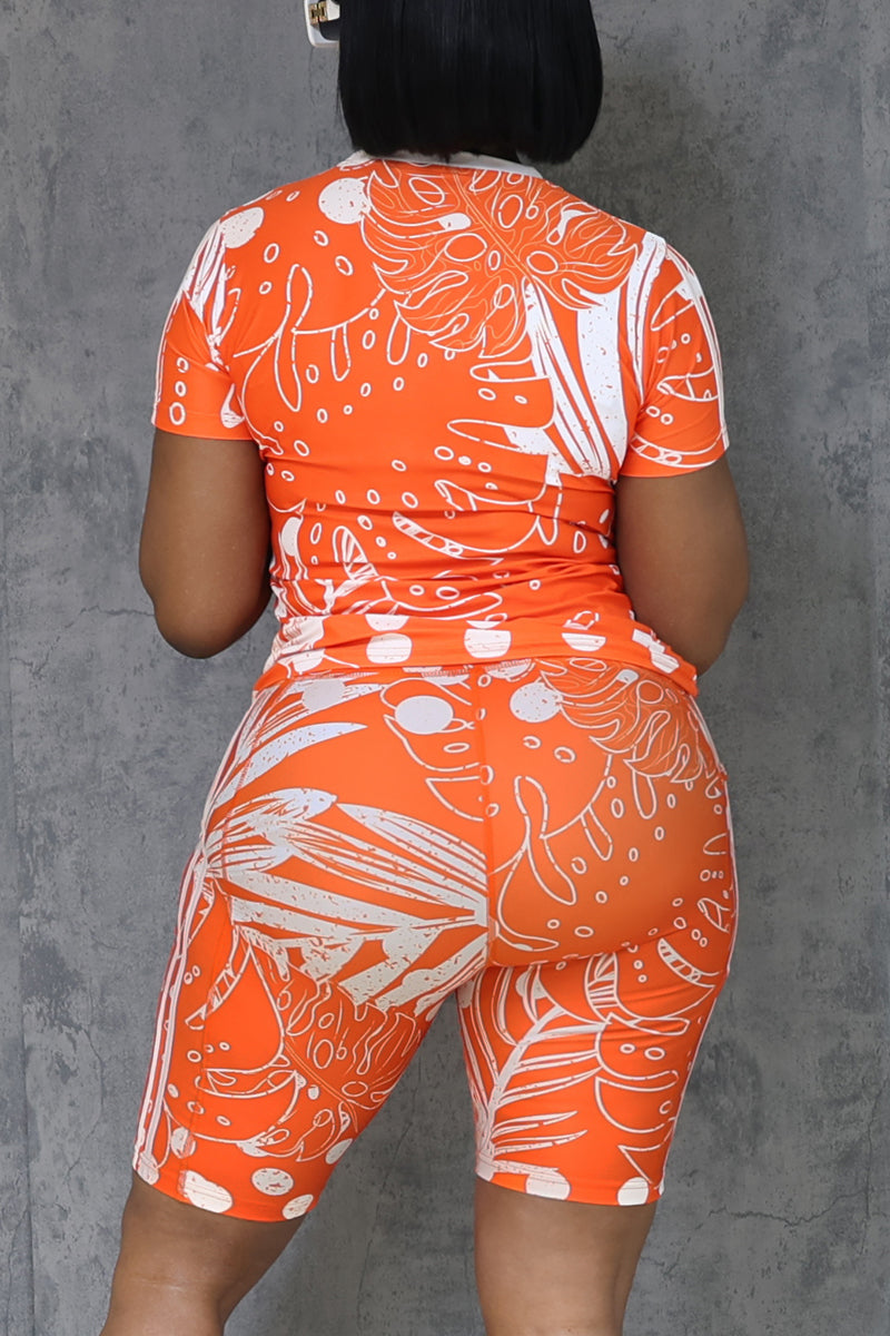 A person with short black hair stands facing a plain gray textured wall, wearing the Orange Summer T-Shirt and matching shorts made from breathable cotton in an orange and white pattern.