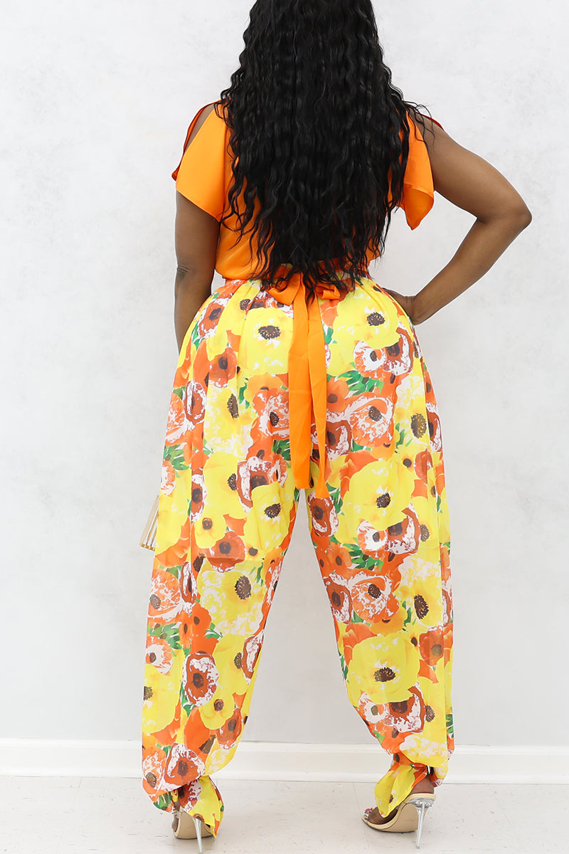 A woman wearing an orange top with yellow and orange floral poppies tie ankle pants and heels.
