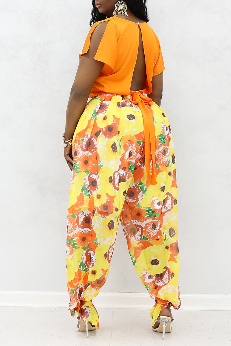 A woman wearing an orange top with  orange and yellow floral poppies tie ankle pants and heels.