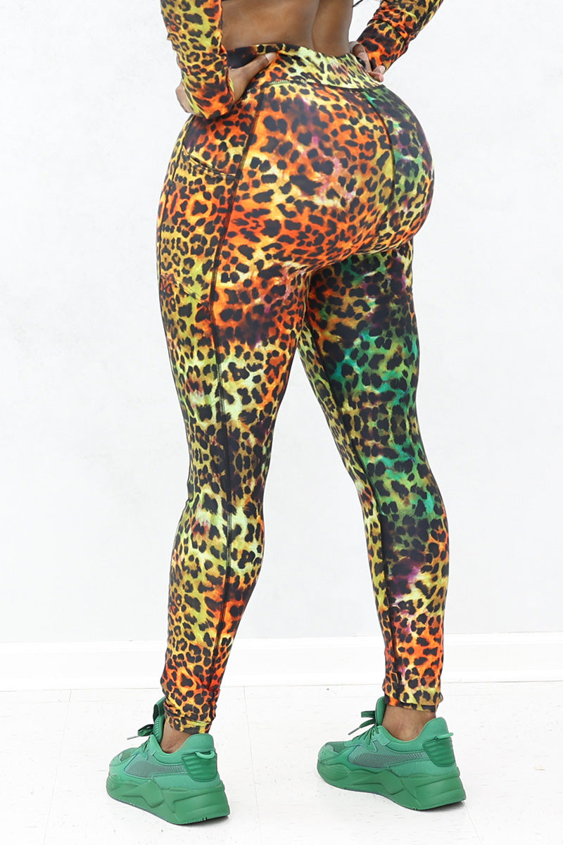 A person faces away from the camera, wearing Safari Tights with a vibrant cheetah print and matching stretchy top, paired with green sneakers. The background is plain and light-colored.