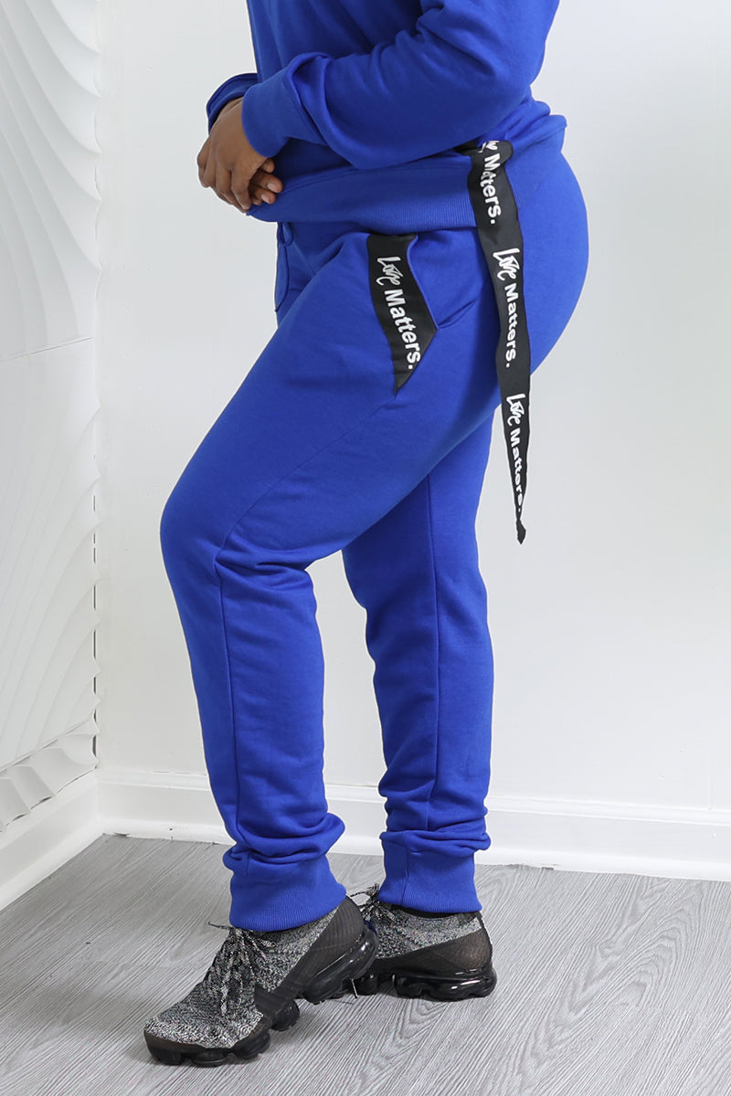 In a room with a white textured wall, the focus is on someone wearing Love Matters Joggers—a blue tracksuit with black ribbon details and gray-black sneakers. The gray wood flooring enhances their sporty vibe.
