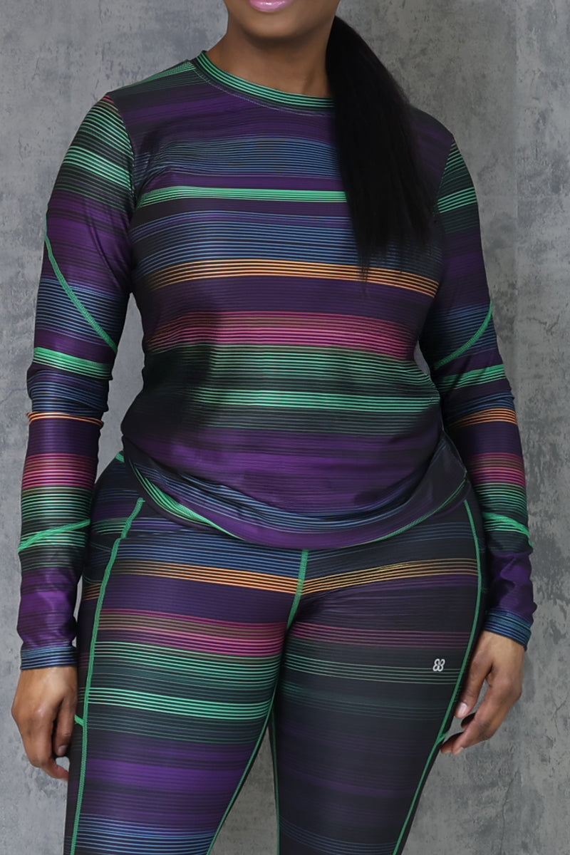 A person stands against a gray textured wall, wearing the Sidelines Long Sleeve T-Shirt in Multi with matching leggings featuring contrast stitching and purple, green, and orange stripes. Their face is partially out of frame.