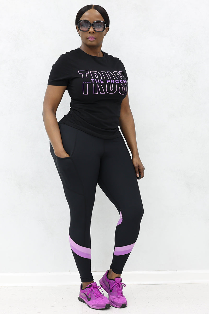 A girl wearing black glasses, a half-sleeve T-shirt with the text "Trust the Process" written on it, black tights and pink shoes.