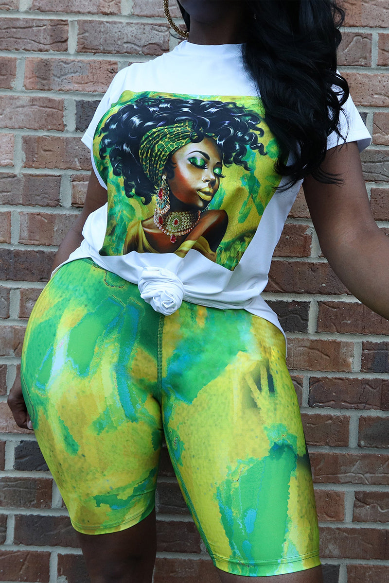A woman wearing a white T-shirt with a black woman's image printed in a square and green shorts is standing against a brick wall.