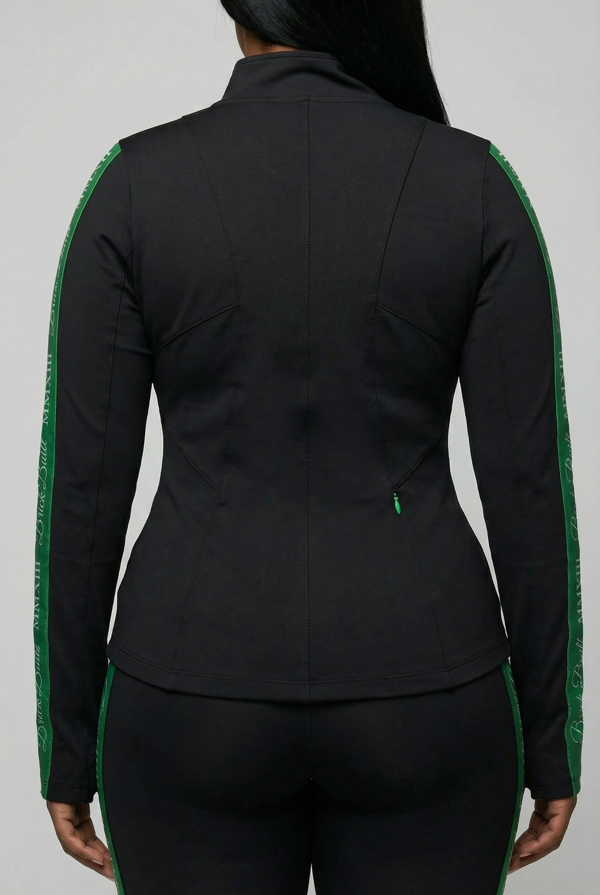 A person is seen from the back wearing the Brick Built 2013 Jacket—fitted in black with green stripes, small green zippers, and bold green sleeve text—contrasting against a plain light gray background.