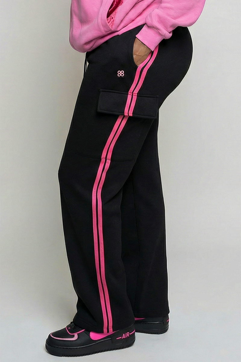 Someone wearing Two Stripe Cargo Pants in black with pink side stripes and a Brick Built logo, paired with black sneakers with pink accents and a pink hoodie, stands against a plain background with their right hand in the pants pocket.