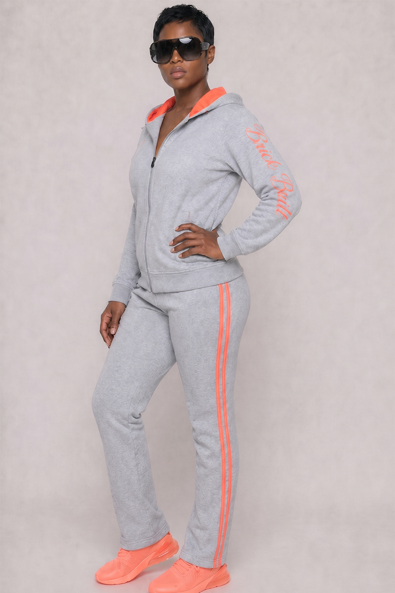 Model wears Grayline Sweatpants (SAMPLE), a grey tracksuit with orange stripes and "Bride Ride" on the sleeve. Paired with sunglasses and orange athletic shoes, they display Brick Built style against a plain light background.