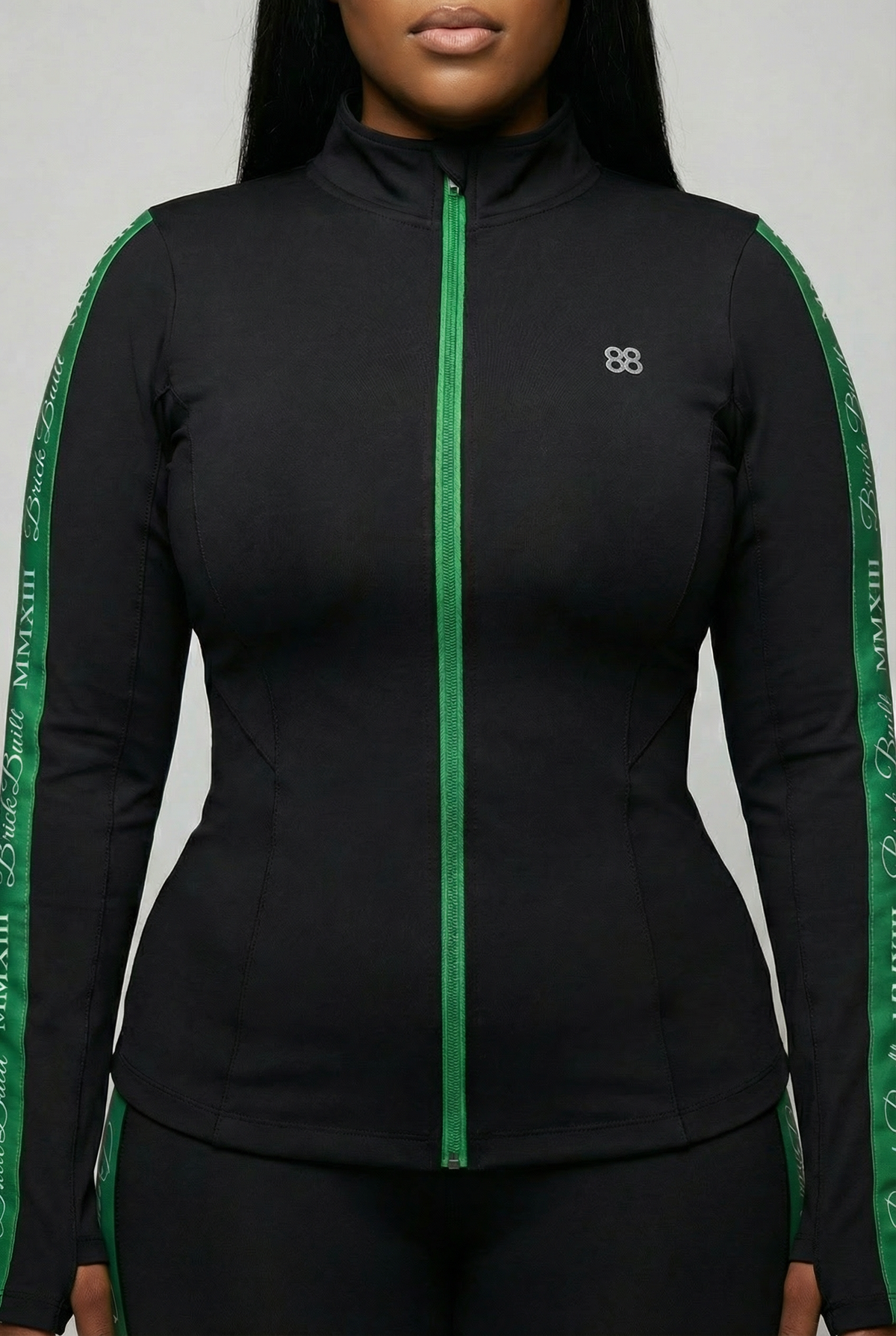 A woman models the Brick Built 2013 Jacket—a fitted black jacket with green stripes, a green zipper, thumbhole sleeves, and a small white chest logo. She stands against a plain background, her face partially out of frame.