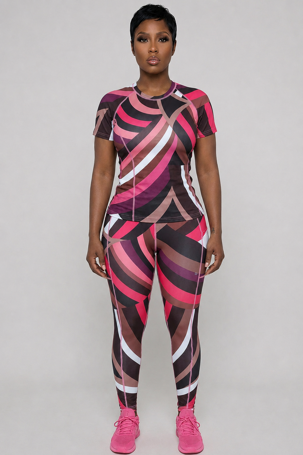 A person faces forward wearing the Sable Tshirt in a relaxed fit, featuring lightweight, breathable fabric with bold curved pink, white, brown, and black patterns, paired with pink athletic shoes against a plain light gray background.