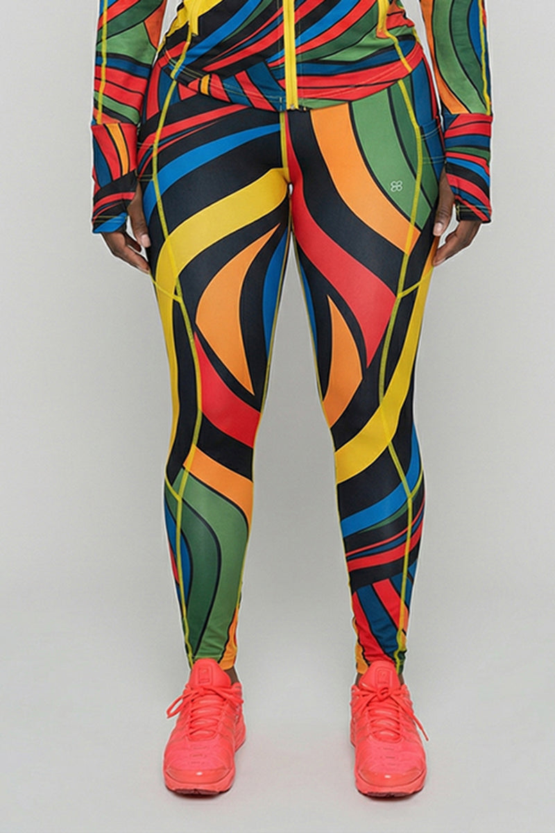 A person wearing Color Riot Tights with a matching abstract-patterned jacket and bright pink athletic shoes stands facing forward against a plain, light background.