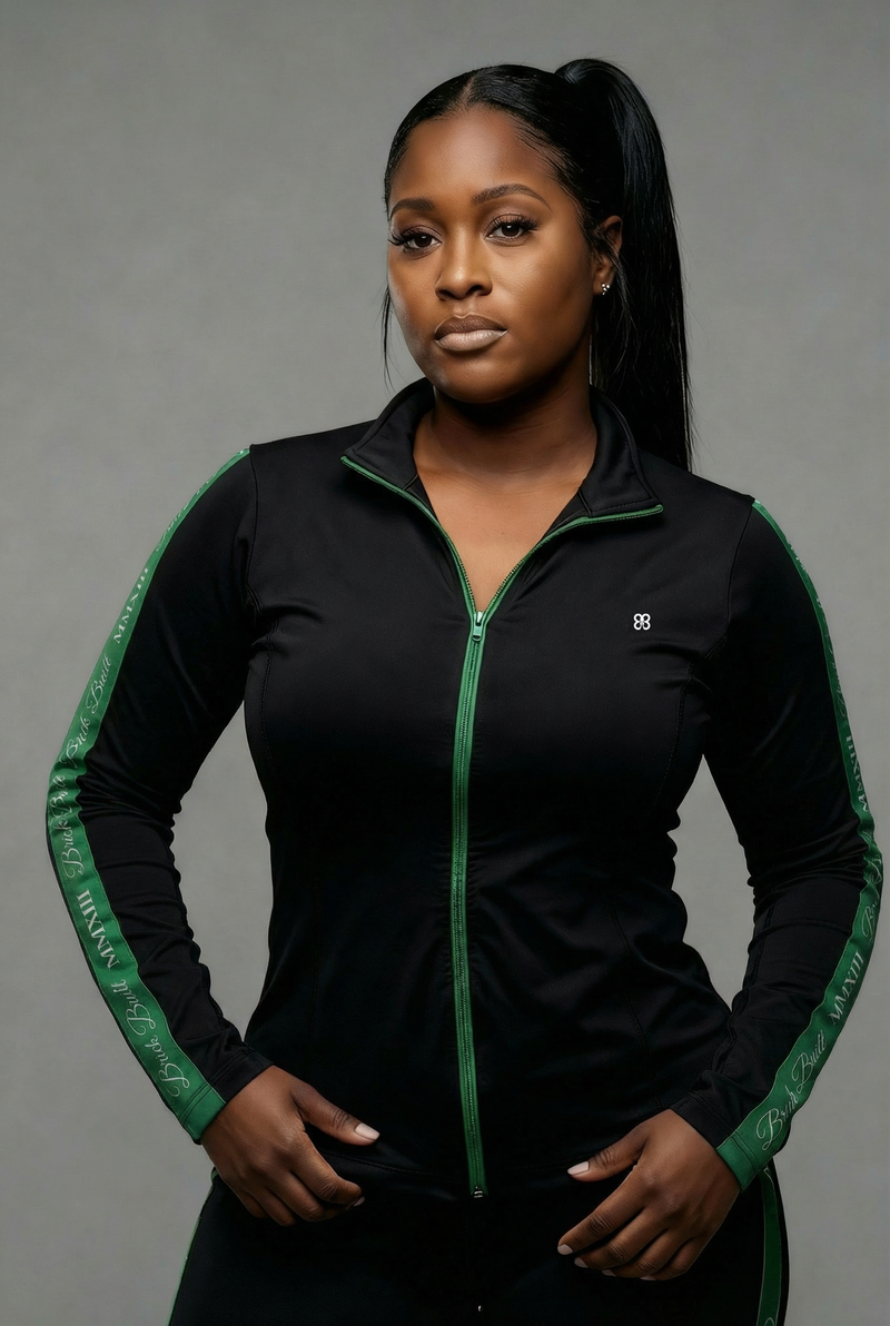 A woman with long dark hair in a ponytail wears the Brick Built 2013 Jacket in Black-Green, featuring thumbhole sleeves, standing against a plain gray background with a neutral expression and her hands at the bottom of the jacket.