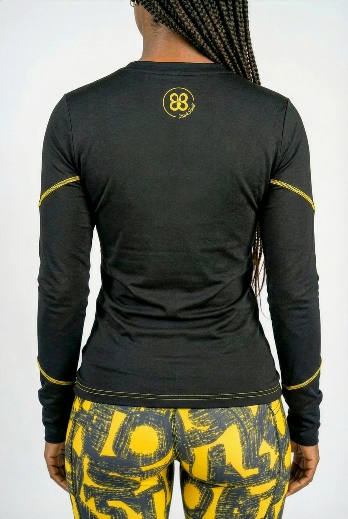 A person with long braided hair is shown from behind, wearing the Black & Yellow Tshirt featuring yellow stitching and a small yellow logo, paired with moisture-wicking yellow and black patterned leggings.