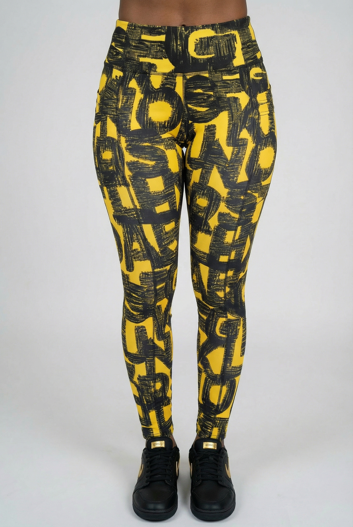 A person stands against a plain background, wearing Black & Yellow Tights featuring a black abstract letter pattern and black sneakers. The waist-down image showcases the stretchy leggings' bold design.