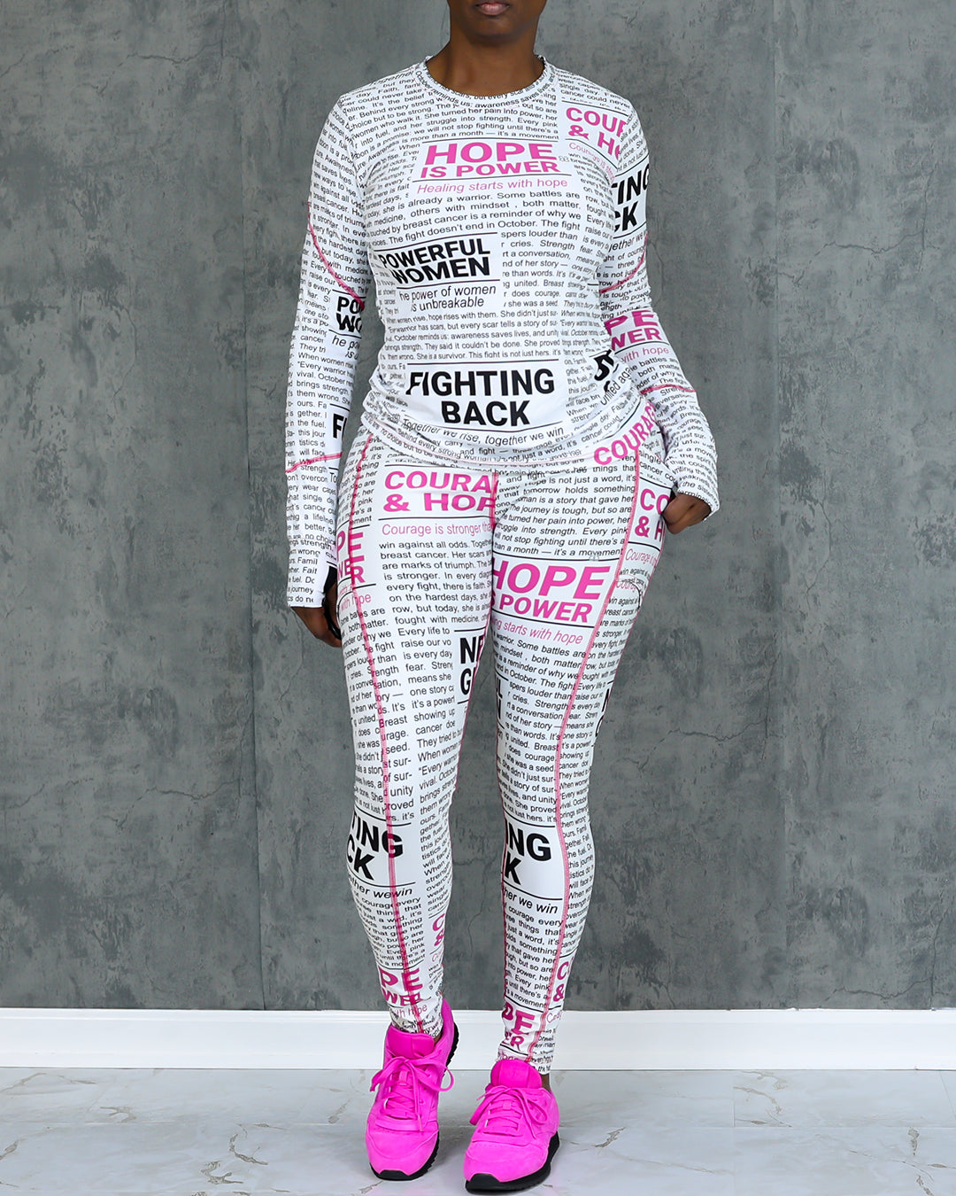 A person stands against a gray textured wall wearing Pink News Tights, made from white 4-way stretch compression fabric with black and pink motivational text like HOPE IS POWER and FIGHTING BACK, paired with bright pink sneakers.