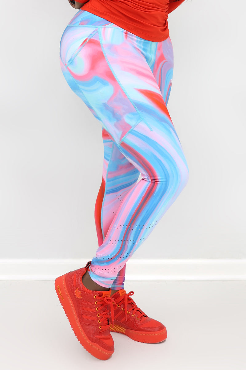 A woman wearing marbled tights in vibrant colors, featuring a drawstring waistband and laser perforations at the ankles. The tights have deep side pockets and a zippered pocket on the back, showcasing a blend of comfort and style ideal for workouts or everyday wear.