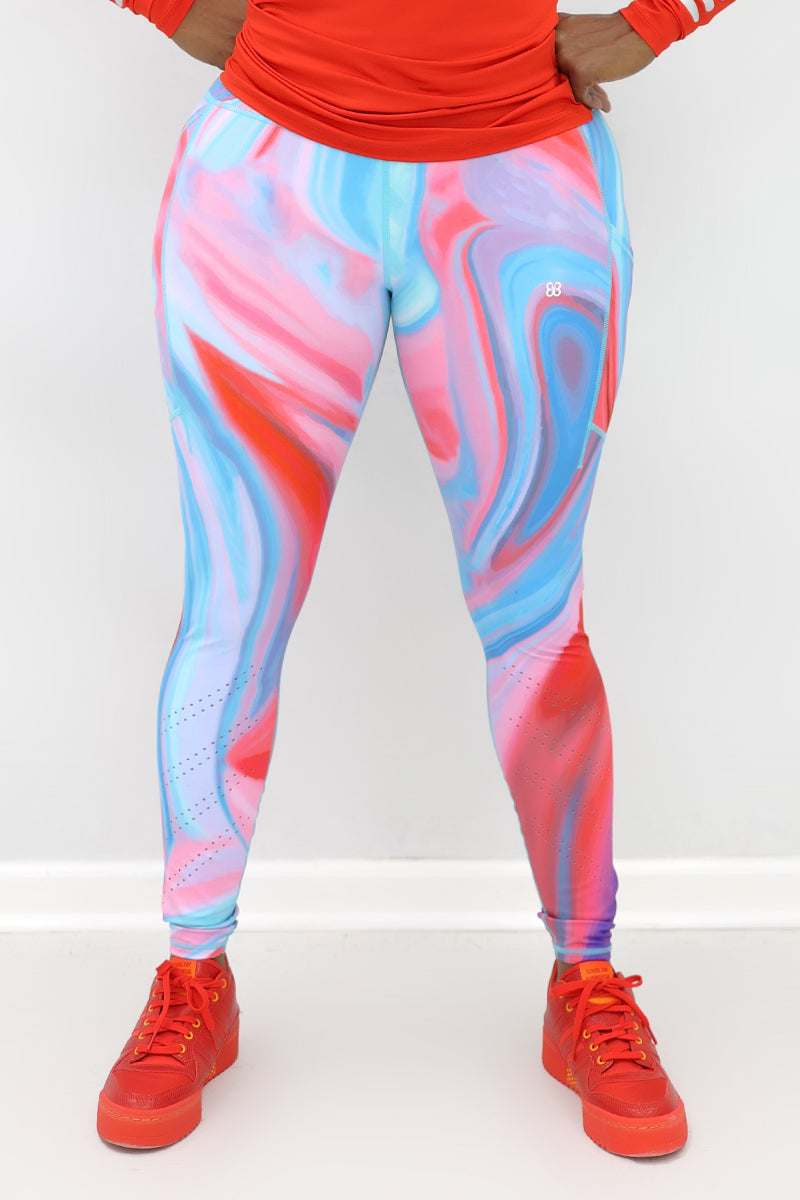 A woman wearing marbled tights in vibrant colors, featuring a drawstring waistband and laser perforations at the ankles. The tights have deep side pockets and a zippered pocket on the back, showcasing a blend of comfort and style ideal for workouts or everyday wear.