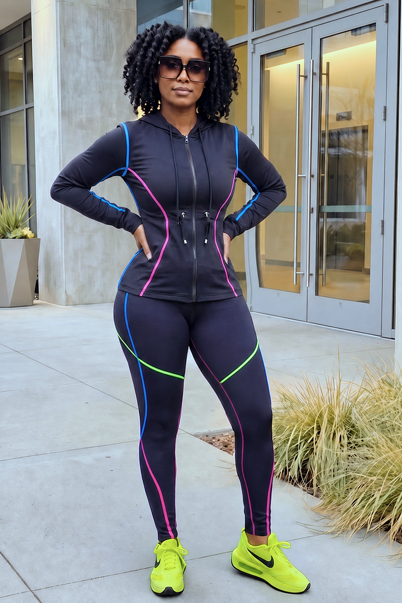 A woman stands confidently outside a modern building with glass doors and concrete walls, wearing the Multi-Stitching Long Sleeve Jacket 1st Edition—a black, stretchy jacket with colorful piping and a full front zipper—paired with neon yellow Nike sneakers.