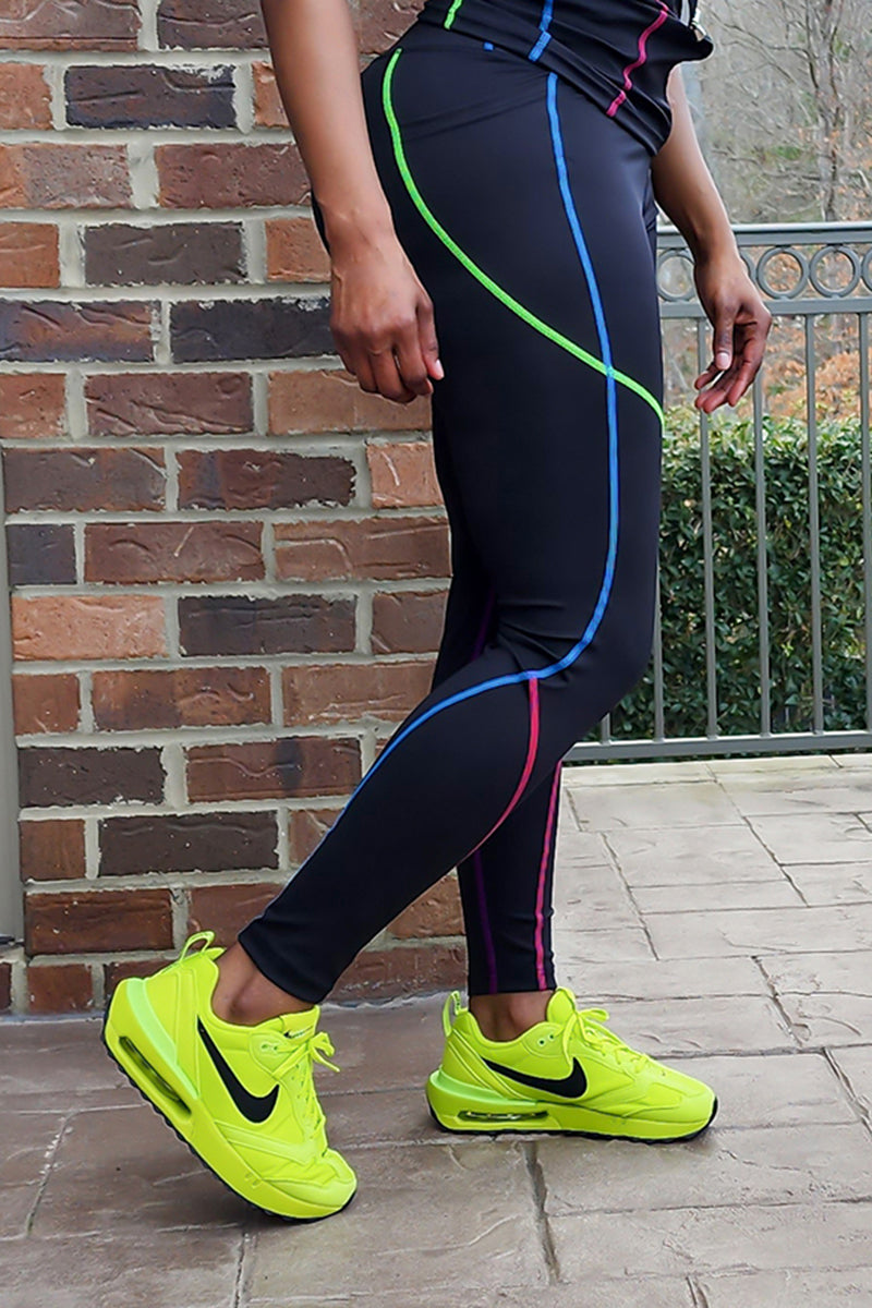 Outdoors on a tiled patio, a person wears Multi-Stitching Tights 1st Edition—black pants with colorful curved lines—and bright yellow Nike Air Max sneakers. A brick wall and metal railing are visible in the background.