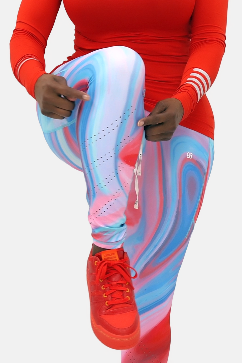 A woman wearing marbled tights in vibrant colors, featuring a drawstring waistband and laser perforations at the ankles. The tights have deep side pockets and a zippered pocket on the back, showcasing a blend of comfort and style ideal for workouts or everyday wear.