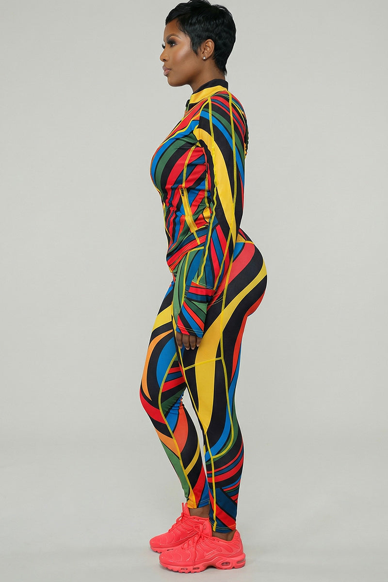 A person stands in profile wearing the Color Riot Jacket, a colorful, form-fitting piece with a geometric pattern and 4-way stretch, paired with bright pink athletic shoes against a plain background.