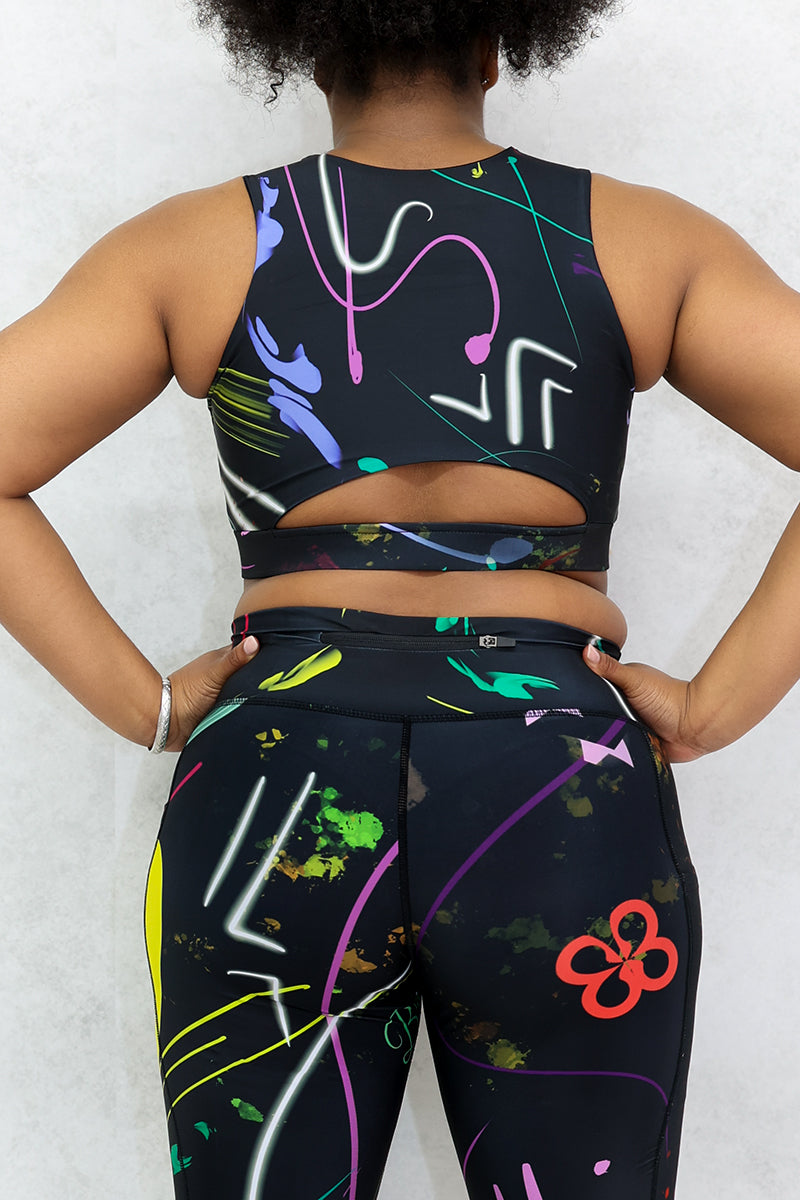 A woman wearing multi-colored Space Girl Tights with a glow effect, featuring the Brick-Built logo, pockets, and a zippered pocket on the back of the waistband, perfect for stylish workouts.