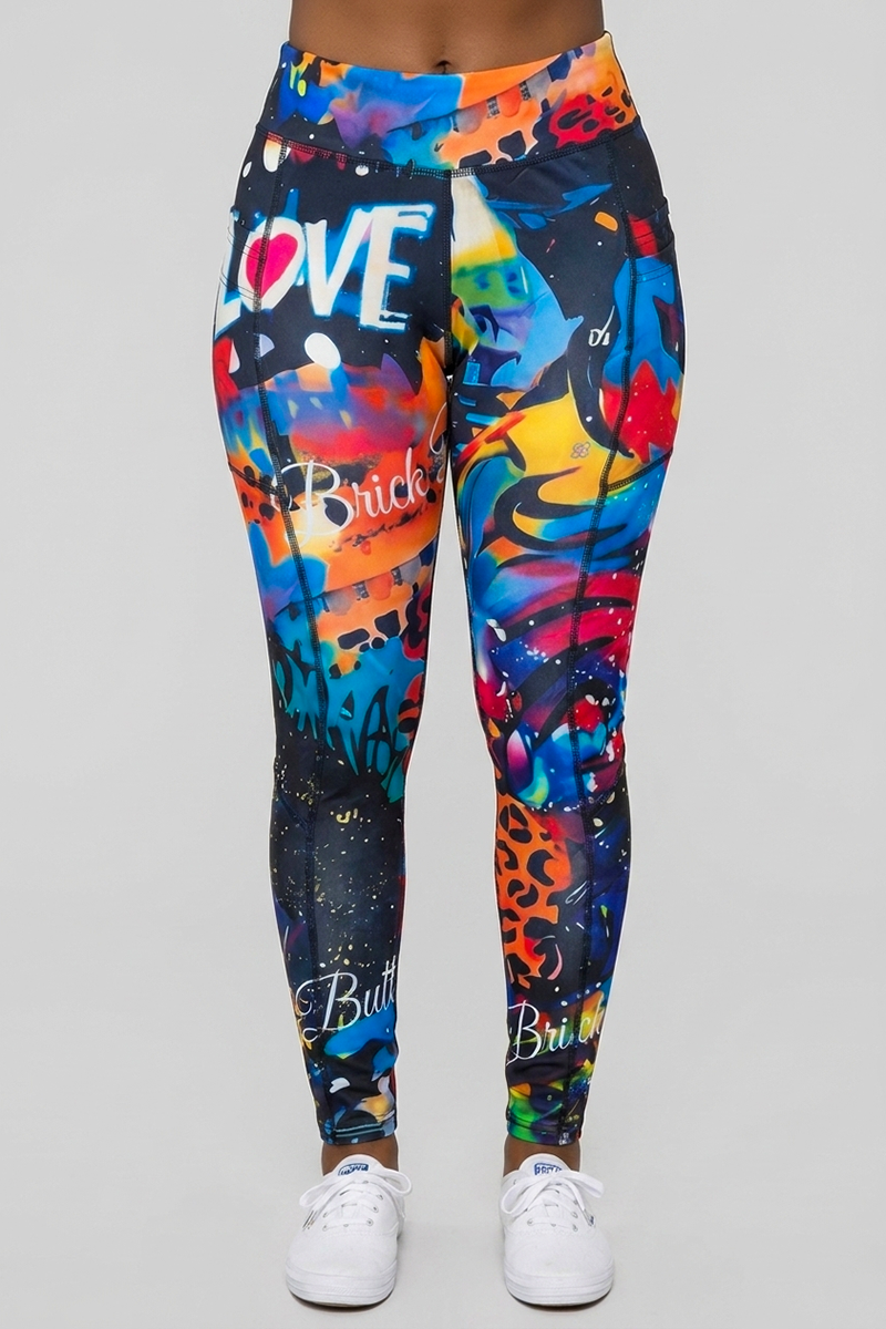 Wearing the Art Code Tights in Multi—a pair of graffiti-inspired leggings made from stretchy polyester spandex with a comfortable 4-way stretch fit and bold abstract patterns in orange, blue, and yellow featuring text including "LOVE"—the person stands against a plain gray background paired with white sneakers.