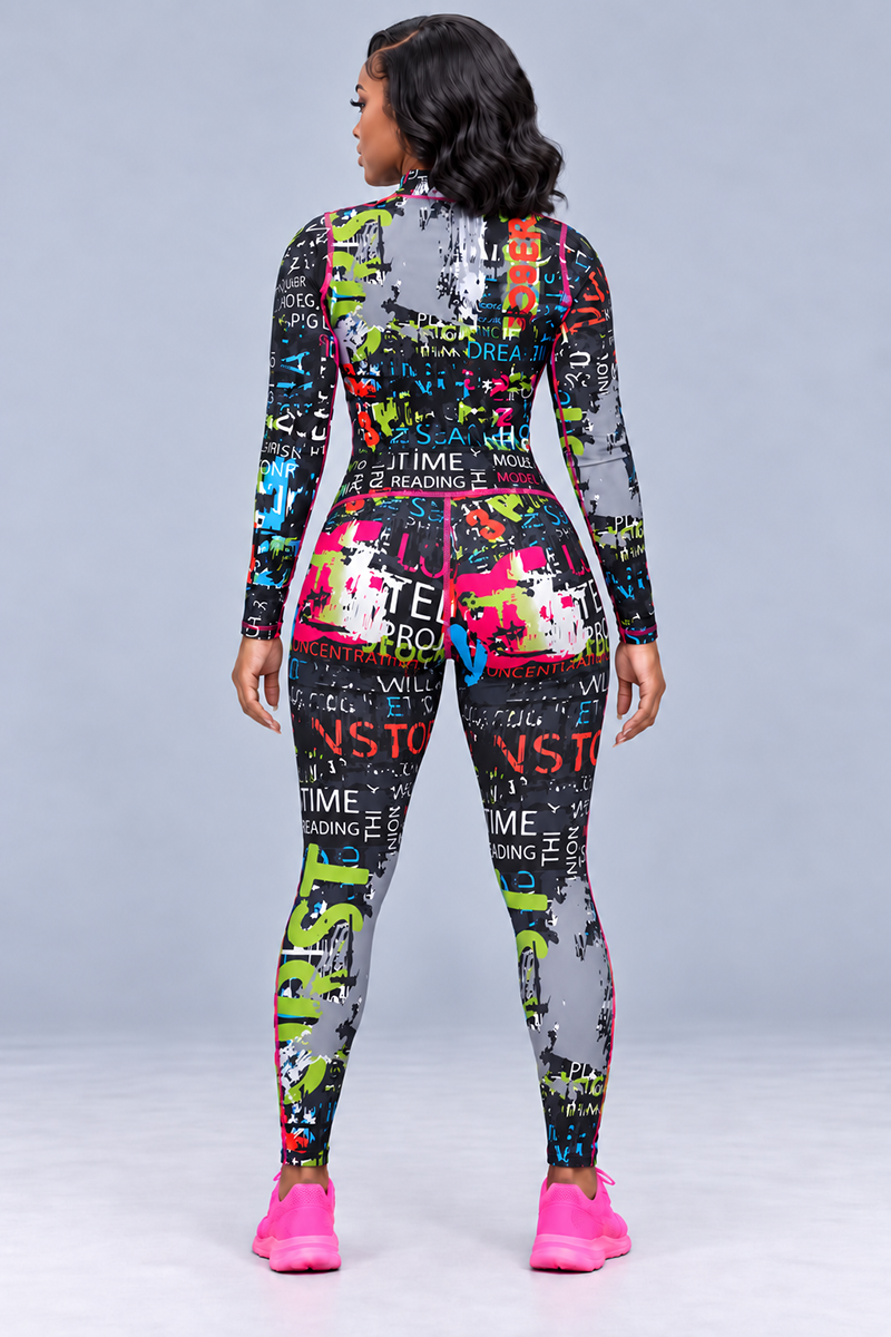 A woman stands facing away, wearing the Electric Letters Half-Zip Pullover—a colorful, long-sleeve top crafted from polyester spandex for 4-way stretch comfort and adorned with graffiti-style text and patterns. She pairs the look with bright pink sneakers, all set against a plain light gray background.