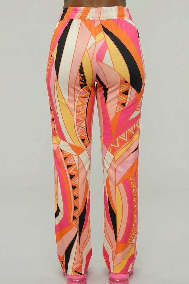 A person stands with their back to the camera, modeling Vanta Pants—brightly colored stretchy pants featuring a geometric pattern in pink, orange, peach, black, and white—against a plain gray background.