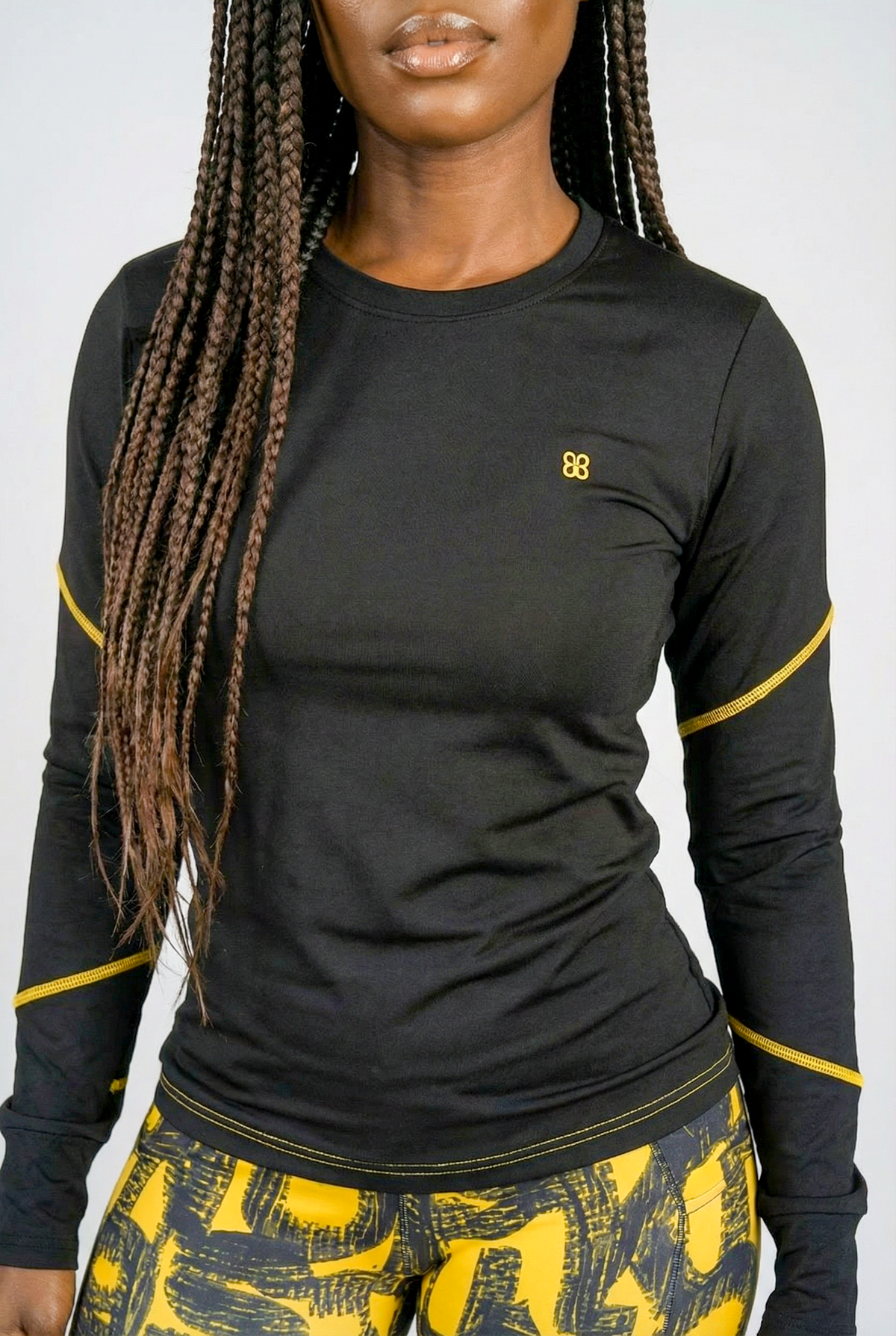 A woman with long braids wears the Black & Yellow Tshirt—a fitted black and yellow top—paired with matching athletic leggings. She stands against a plain light background.