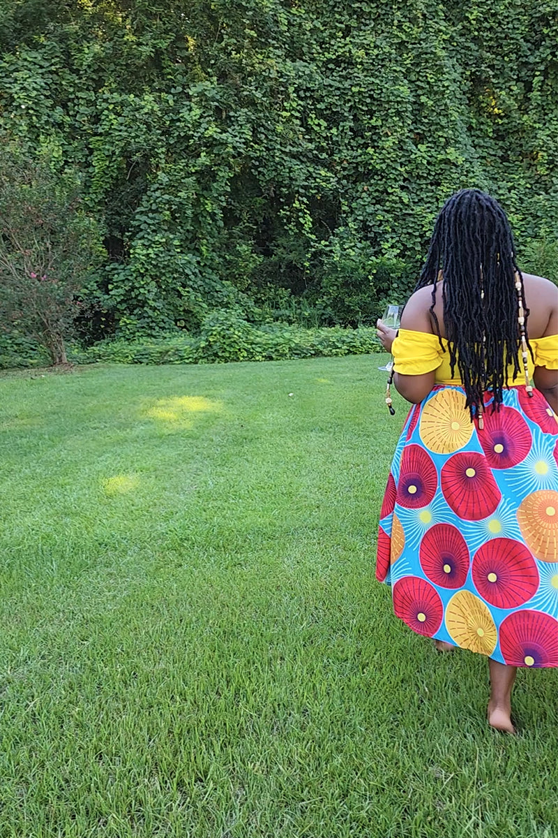 A woman with long braided hair wearing an off-shoulder yellow top and the Red Sun Knee Skirt, walks barefoot on grass with a wine glass in hand. Lush green foliage forms the background.