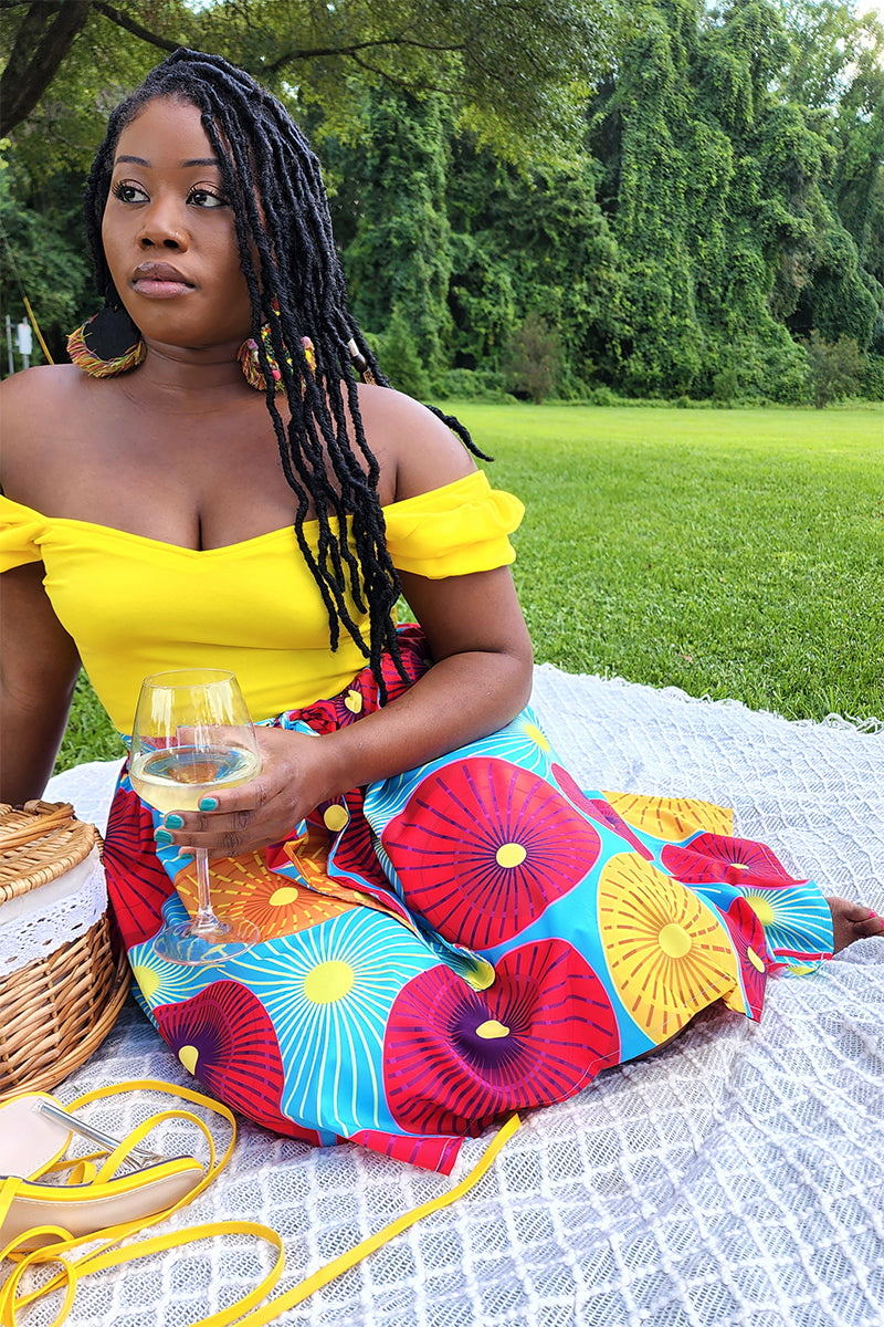 A woman in a yellow off-shoulder top and the Red Sun Knee Skirt sits on a picnic blanket outdoors, holding a glass of white wine, with sandals and a wicker basket beside her on the grass.