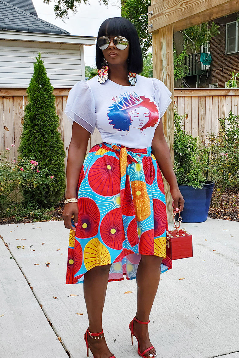 A woman stands outdoors wearing a white graphic T-shirt, the Red Sun Knee Skirt with vibrant blue, red, and yellow patterns, red heels, large earrings, sunglasses, and holding a small red handbag.